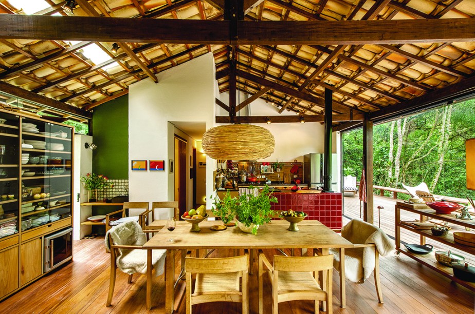 De madeira teca, a mesa e as poltronas dinamarquesas (Casual) re&uacute;nem os amigos e tamb&eacute;m d&atilde;o apoio ao casal quando traz trabalho de S&atilde;o Paulo. Ao fundo, o fog&atilde;o a lenha (Oficina Victorello) foi revestido de azulejos especiais da Antigua, lembrando o vermelh&atilde;o dos fog&otilde;es mineiros. Projeto de Vania Chene.