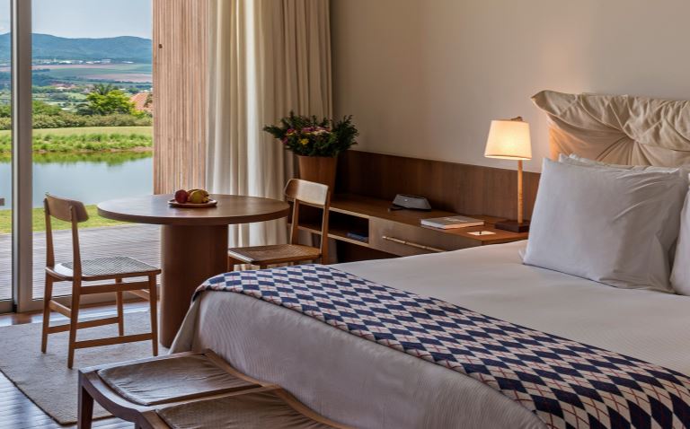 Quarto do hotel Fasano Boa Vista, com cama grande, mesa e duas cadeiras e vista para um lago e montanhas
