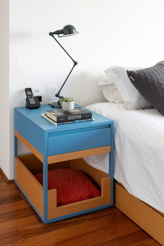 Mesa de cabeceira azul e marro, com a parte inferior com uma almofada vermelha para pets. Acima dela, dois livros, um vasinho com suculenta, uma luminária preta e um telefone sem-fio.