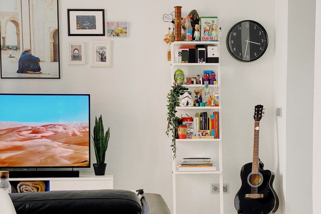 Sala de paredes brancas com quadros pendurados. Relógio de parede e violão à direita. Estante com livros, gadgets e bonecos branca no centro. Televisão no canto inferior esquerdo