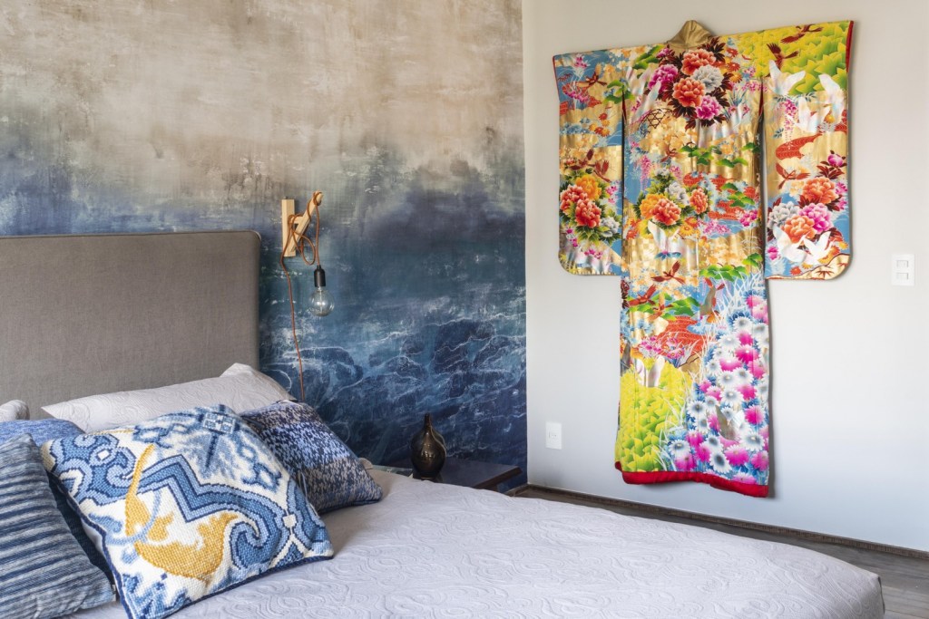 Cama com travesseiros estampados de azul e parede pintada com tons de azul a simular ondas. Um quimono colorido com flores e aves pendurado na parede.