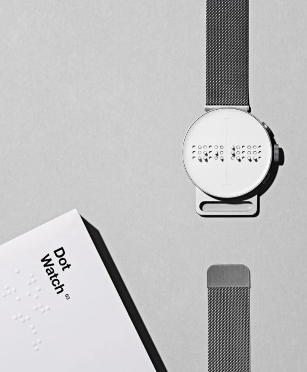 Dot Watch com a pulseira aberta em uma das extremidades em fundo cinza. No canto inferior esquerdo, manual do relógio em braille
