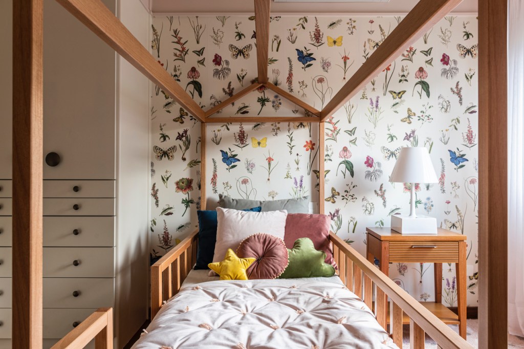 Quarto de criança com cama em madeira em formato de casinha. Travesseiros coloridos, um em formato de estrela, um redondo e um em formato de nuvem. Papel de parede com flores, borboletas e folhas em estilo botânico