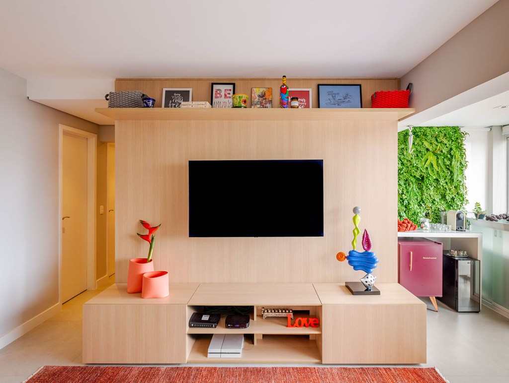 Móvel de madeira que serve como suporte da tv e armário. Prateleira no alto com porta retratos. Vaso laranja salmão. Varanda à direita