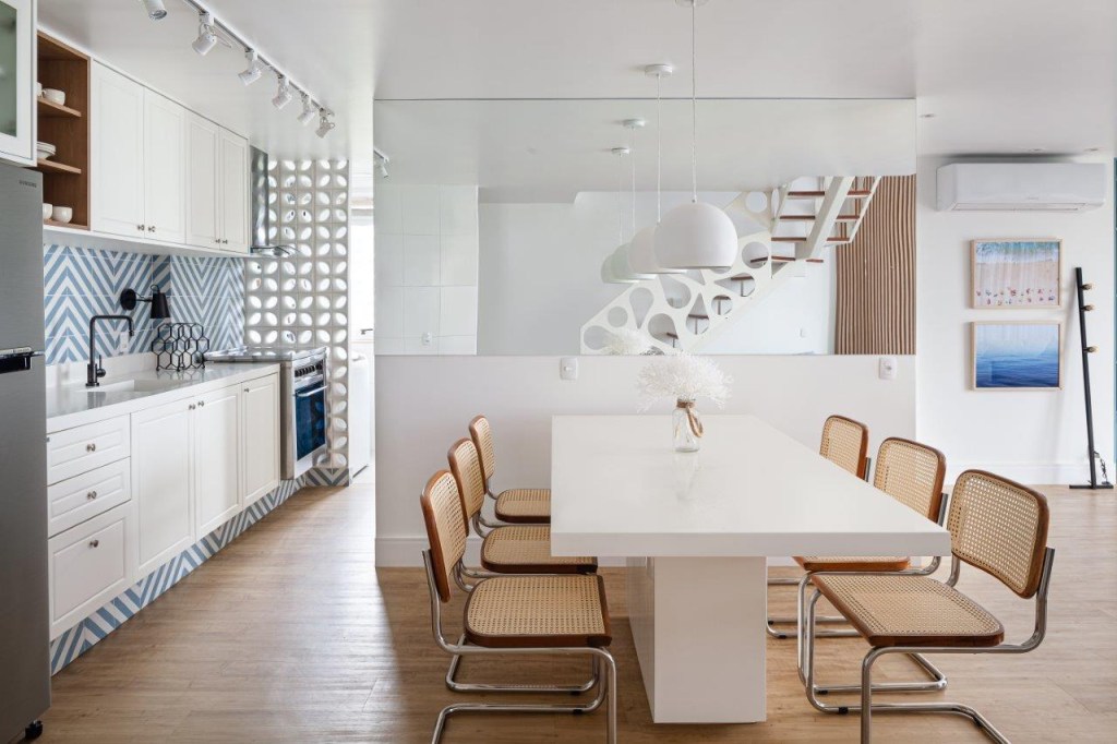Sala de jantar com mesa branca de seis lugares. Cozinha integrada ao fundo na esquerda com parede de azulejos azuis e brancos no padrão listrado. Cobogós brancos ao fundo da cozinha. Escada ao fundo no centro refletido no espelho.