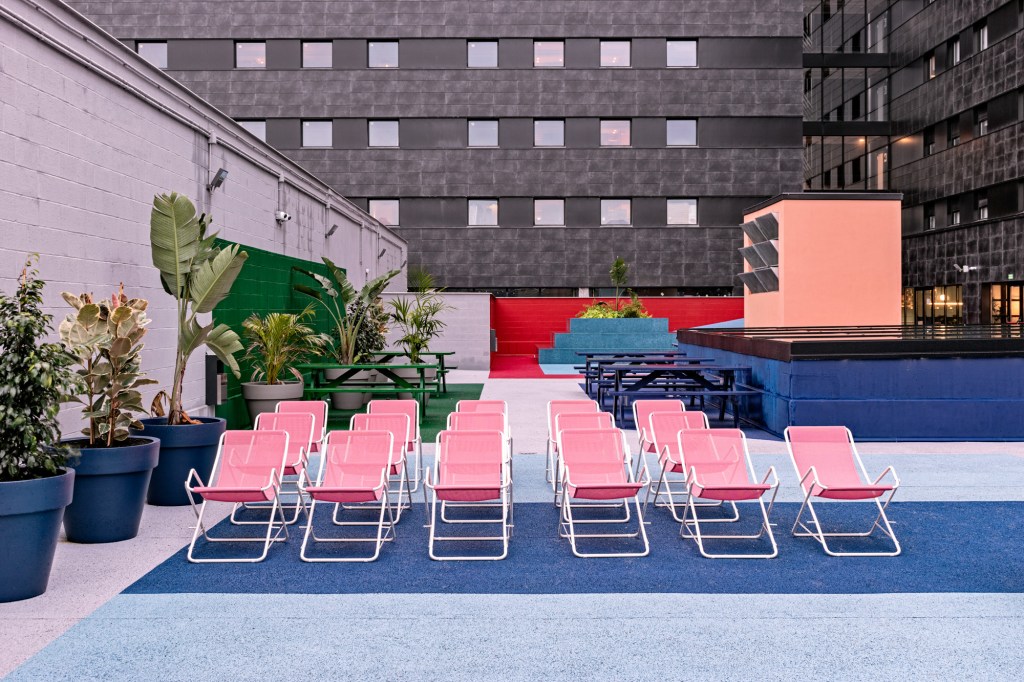Terraço aberto com chão em tons de azul, com cadeiras de praia rosa, paredes cinzas, com parte pintada de ver, com mesas da mesma cor