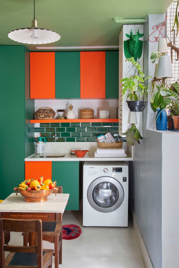 Lavanderia com armários verdes e laranjas. Máquina de lavar encaixada. Plantas penduradas