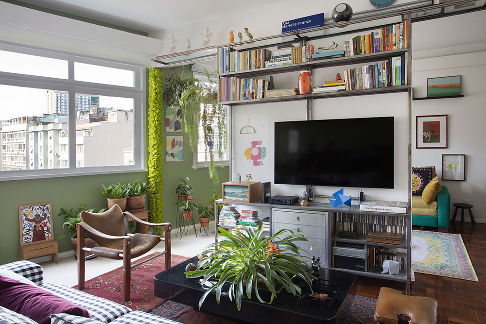 Sala de tv com sofá listrado, móvel cinza com livros e tv. Tapete vermelho. Cadeira de madeira. Plantas ao fundo