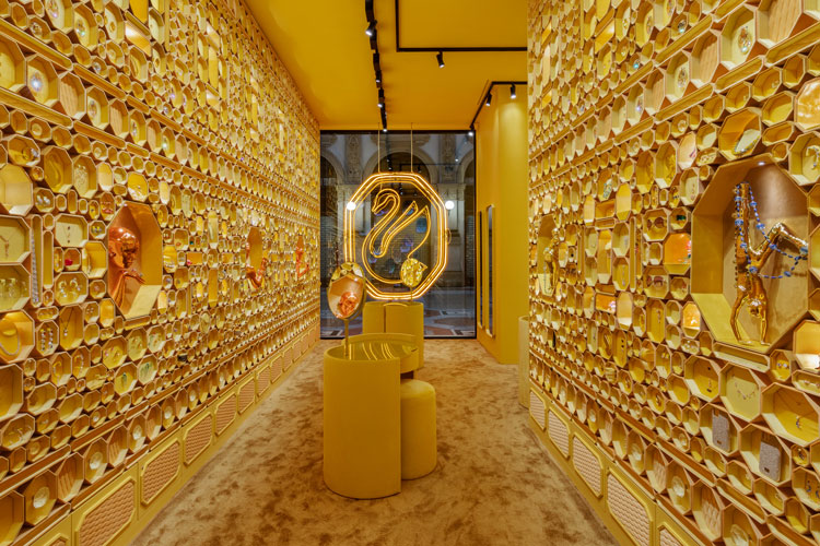 Loja com paredes cobertas por caixas da Swarovski em forma de octógono douradas. Vitrine ao fundo com novo logo de cisne dourado em luz