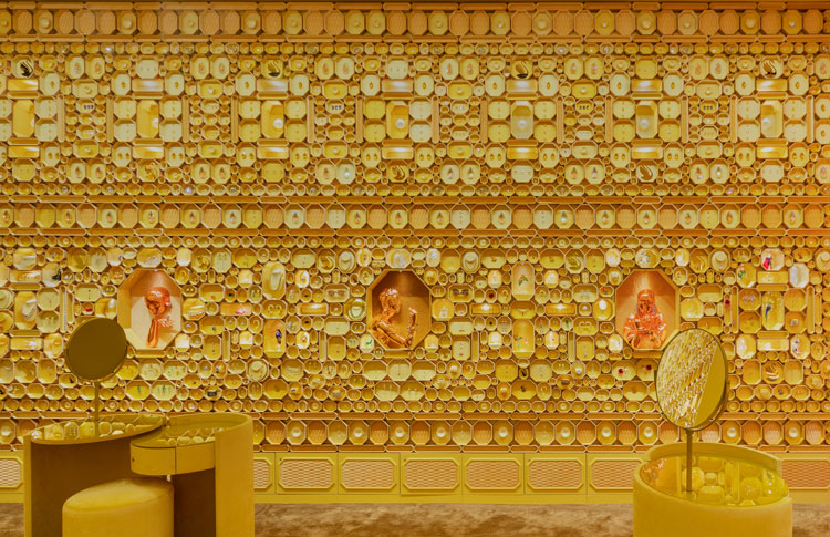 Parede coberta com caixas de diferentes tamanhos douradas. Mesinhas com pufes também amarelos.