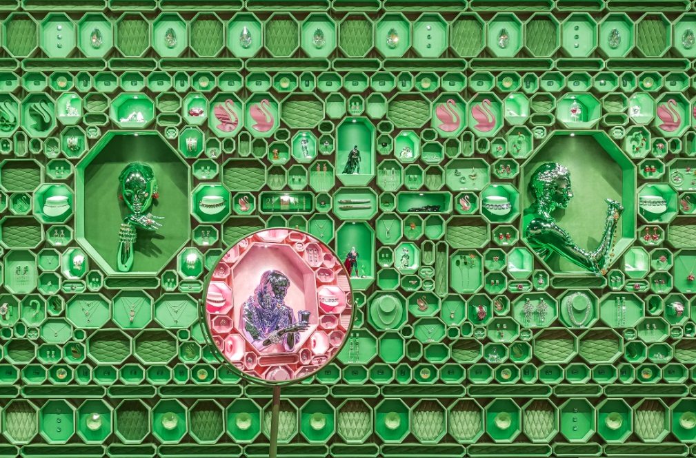 Parede coberta com caixas octogonais verdes. Espelho no canto inferior esquerdo reflete a parede rosa do lado oposto.