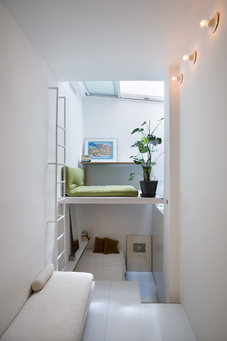 Quarto em dois níveis estreitos. Almofada verde, planta e quadro no andar de cima. Escada branca. No andar de baixo, cama.