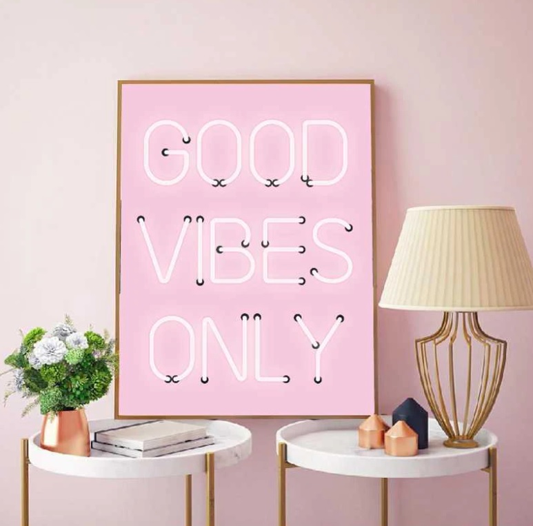 Quadro rosa com letras em neon escrevendo Good vibes only. Duas mesinhas brancas de tampo redondo com abajur e vasinho de flor