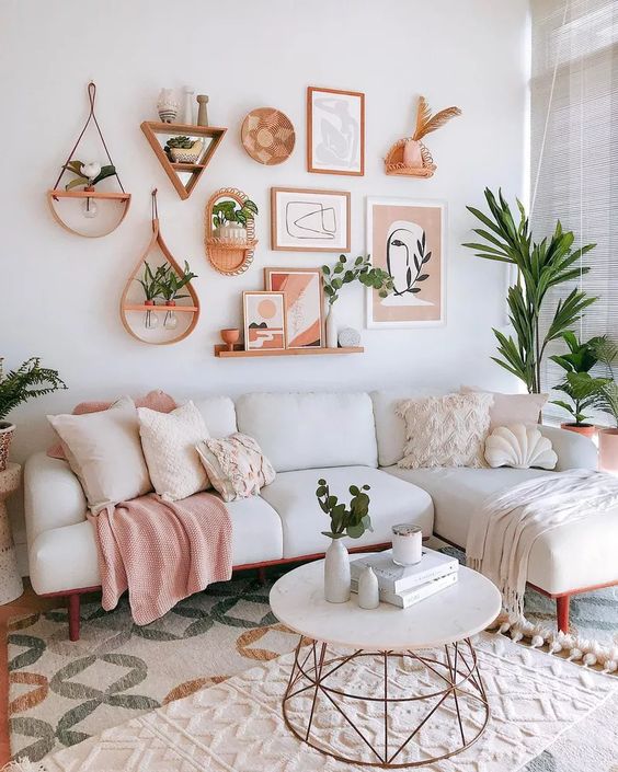 Estar com sofá branco e almofadas brancas e rosa claras. Uma almofada em formato de concha. Na parede, quadros, vasos de plantas e uma prateleira com pequenos quadros