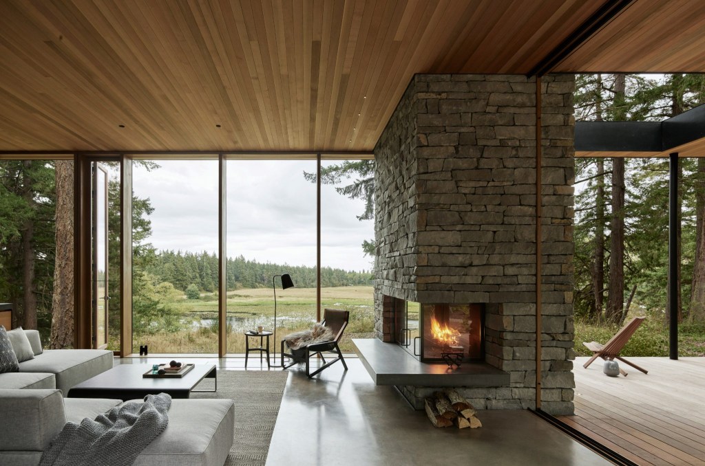 Ambiente calmo com madeira e pedra. Grandes janelas do chão ao teto dão vista para um grande campo aberto