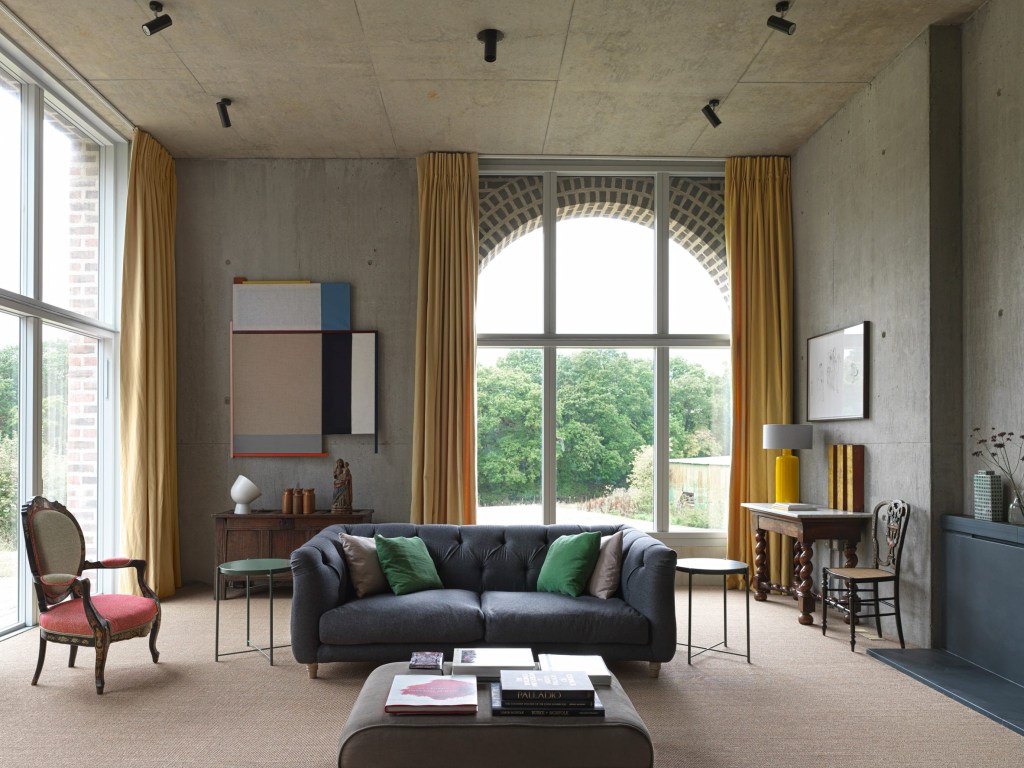 Sala de estar com tetos altos de concreto como um fundo calmo. Uma cadeira de estilo barroco e um banquinho revestido de tecido ajudam a suavizar o espaço.