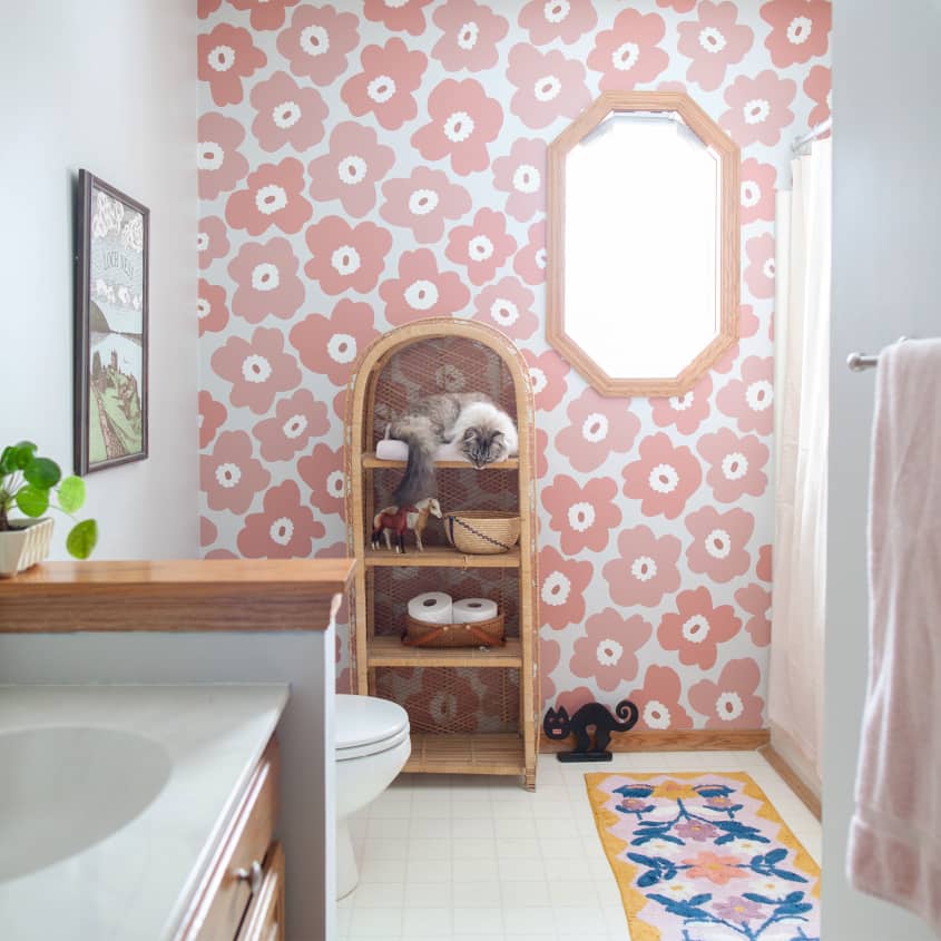 Banheiro grande com papel de parede florido em rosa salmão. Um móvel de madeira com itens de banheiro e um gato. À esquerda, a pia branca e acima do vaso, na parede branca, um quadro emoldurado