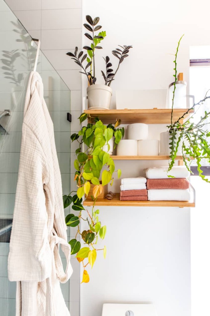 Acima do vaso, três prateleiras, com toalhas, papel higiênico e plantas. Ao lado, um roupão de banho branco pendurado