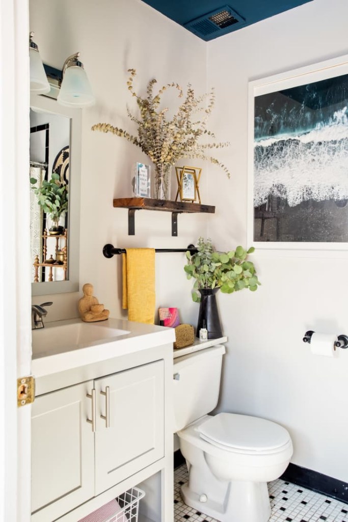Banheiro branco, com prateleiras com porta retratos, vaso e abaixo uma haste de metal com uma toalha amarela pendurada