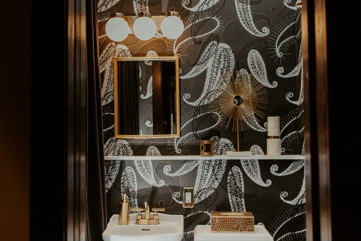Banheiro com papel de parede escuro, com padrão de plantas. Uma prateleira longa decora o ambiente, junto com peças brancas e douradas