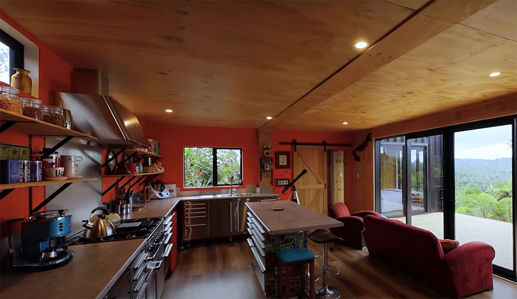 Cozinha integrada com estar. Parede vermelha. Piso e teto revestidos em madeira. Sofá vermelho voltado para a janela