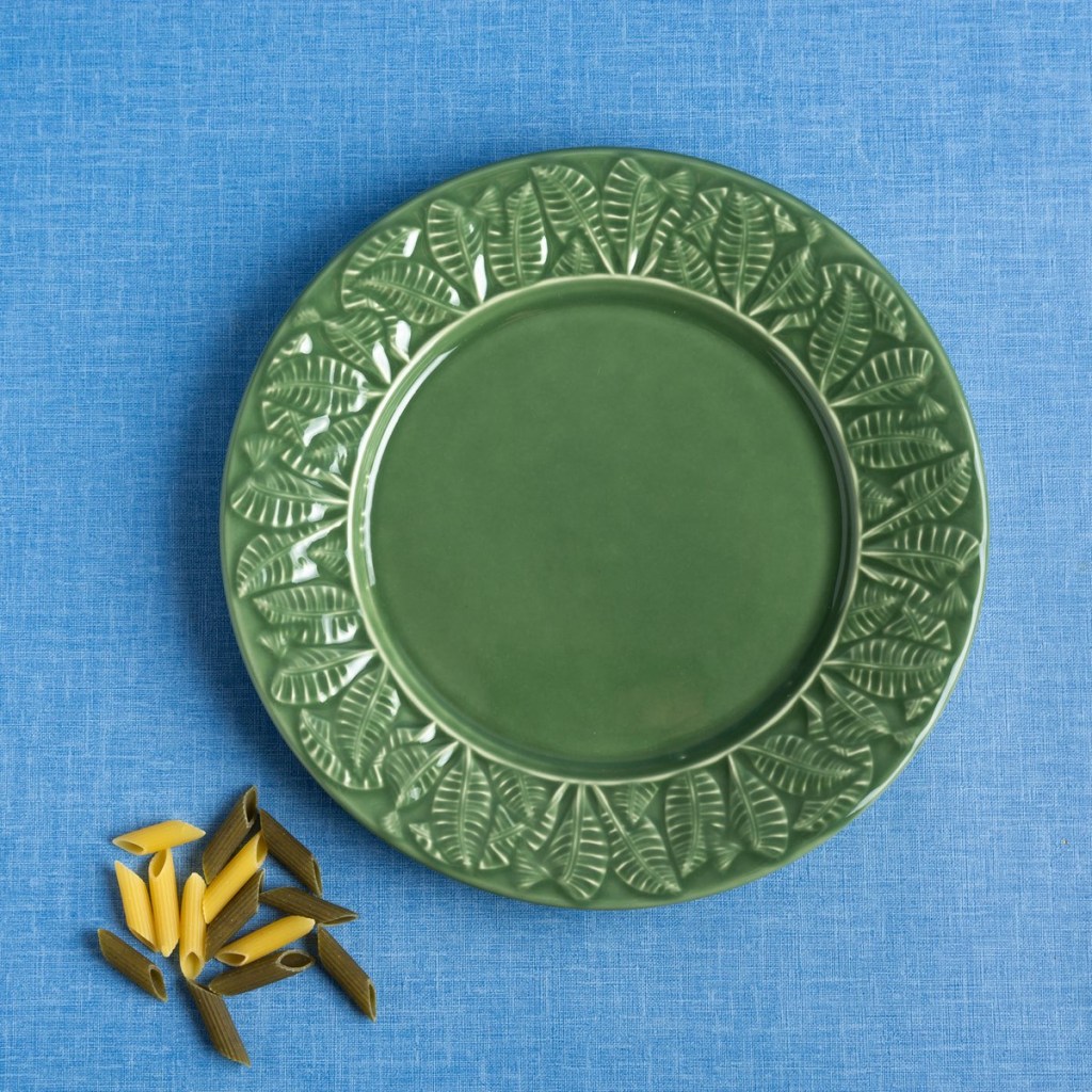Prato raso de cerâmica verde, com desenhos de folhas na borda, em fundo azul mesclado com grãos de macarrão