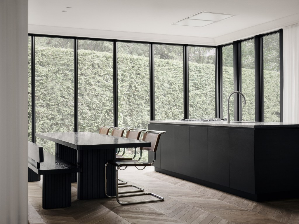 Sala de estar com janelas grandes que cobrem as paredes. Vista para jardim. Mesa e bancada preta. Piso em madeira