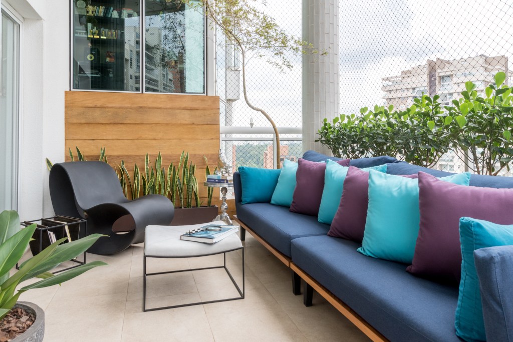 Varanda aberta com porcelanato, mesa de centro branca, com sofá azul com almofadas em azul claro e roxo. Ao lado, com plantas ao fundo, uma cadeira de design completa o ambiente