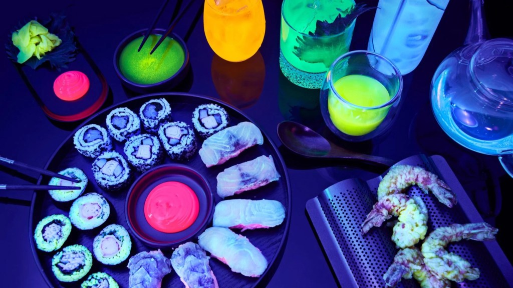 mesa com prato de sushis e camarões empanados com cores neon. drinks ao lado
