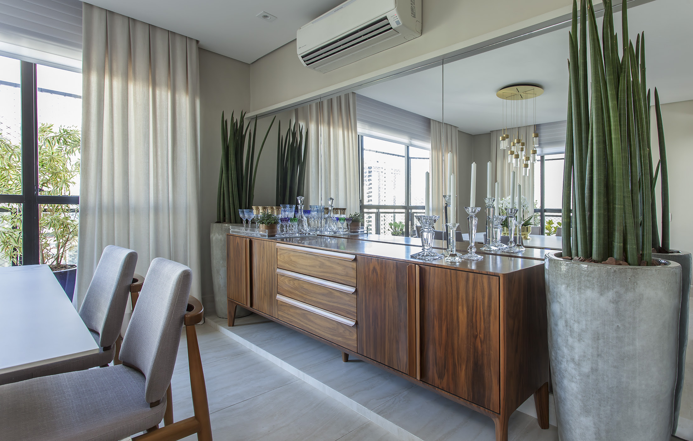 Este buffet em madeira Nogueira acompanha a parede espelhada e &eacute; ladeado por vasos cer&acirc;micos com lan&ccedil;as de S&atilde;o Jorge. Al&eacute;m de sua fun&ccedil;&atilde;o de guardar lou&ccedil;as, revela um d&eacute;cor de personalidade na sala de jantar dos moradores