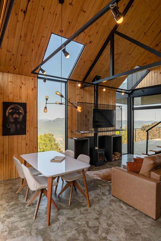 Sala de estar e jantar em cabana de steel frame com revestimento em madeira. Com lareira e abertura em vidro que permite ver a paisagem de montanha.