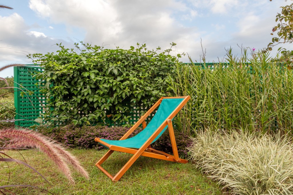 Detalhe de jardim na cobertura da casa, com cadeira de descanso em madeira e lona e plantas com céu ao fundo.