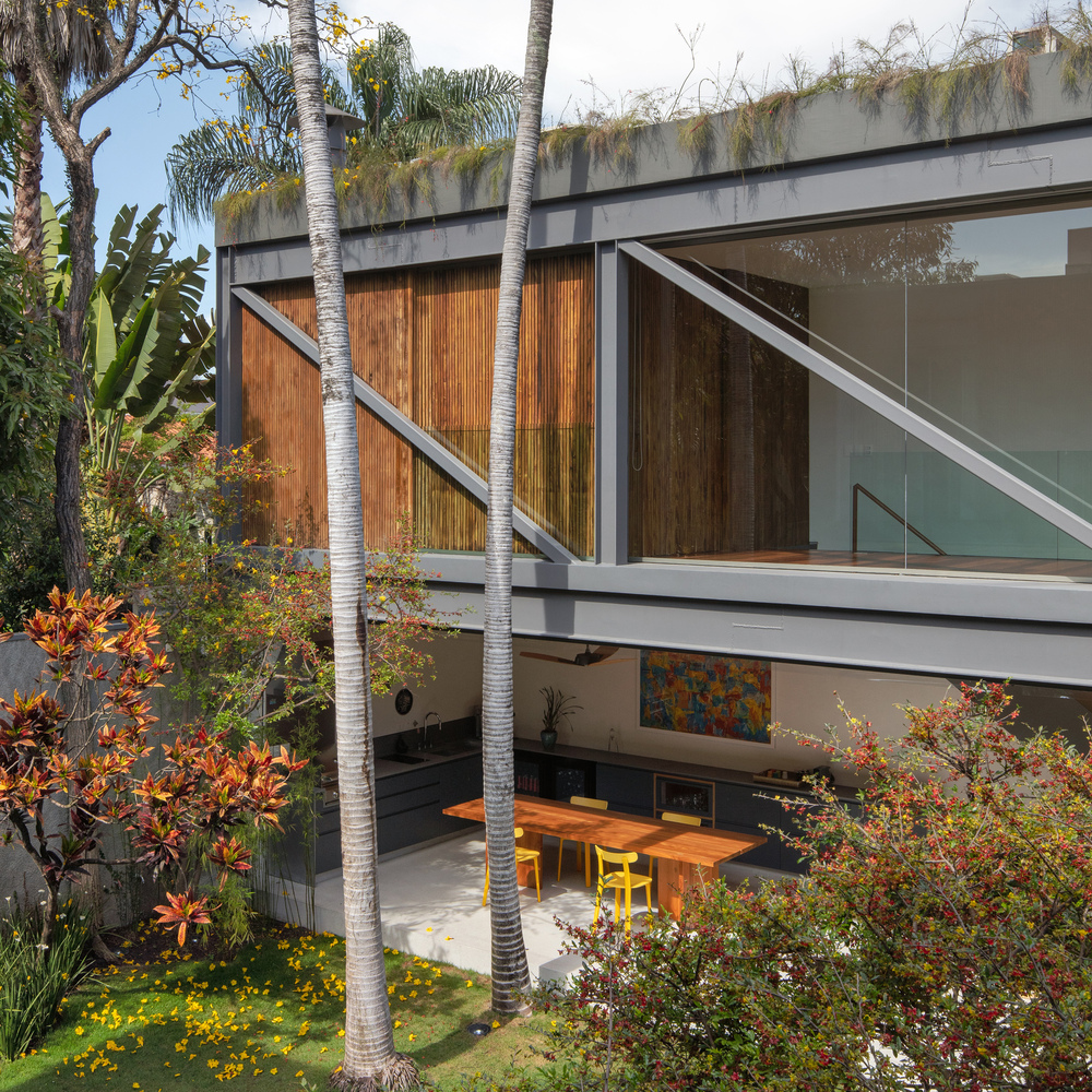 Edícula da casa com piso térreo integrado à área externa e piso superior em fechamento em ripado de madeira e vidro. Dá para ver parte da sala de ginástica. Vegetação na cobertura.