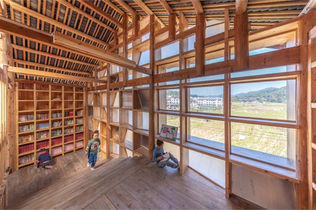 Área interna da biblioteca comunitária construída em madeira. Duas crianças brincam perto da fachada em madeira e policarbonato.