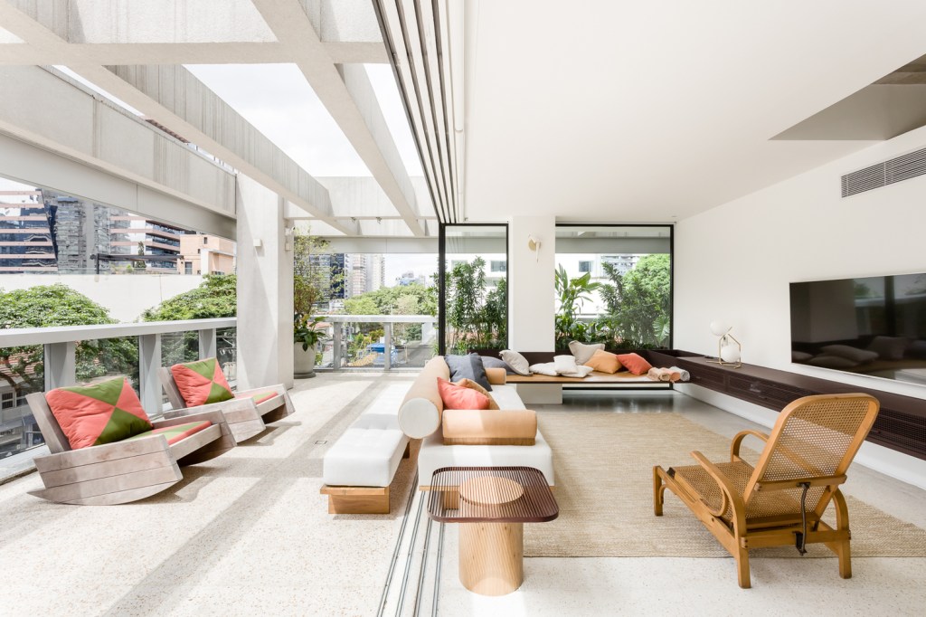 Sala de estar em apartamento tem varanda integrada e coberta com pérgula de vidro. Ambiente claro, com luz natural e móveis de madeira com almofadas coloridas.