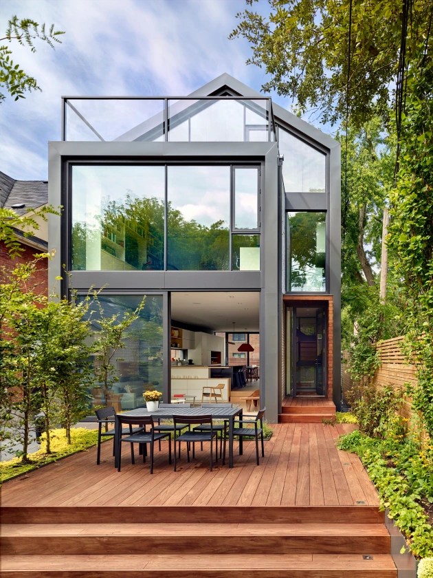 Foto mostra casa em estrutura metálica com deck de madeira que abriga área de refeições com mesa grande e cadeiras, rodeado pelo jardim.