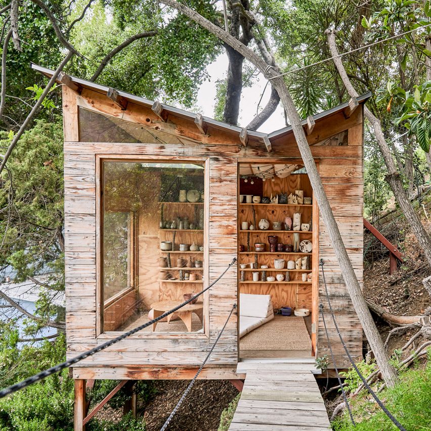 Cabana da madeira sobre palafitas, com aberturas de vidro e prateleiras internas que exibem cerâmicas.