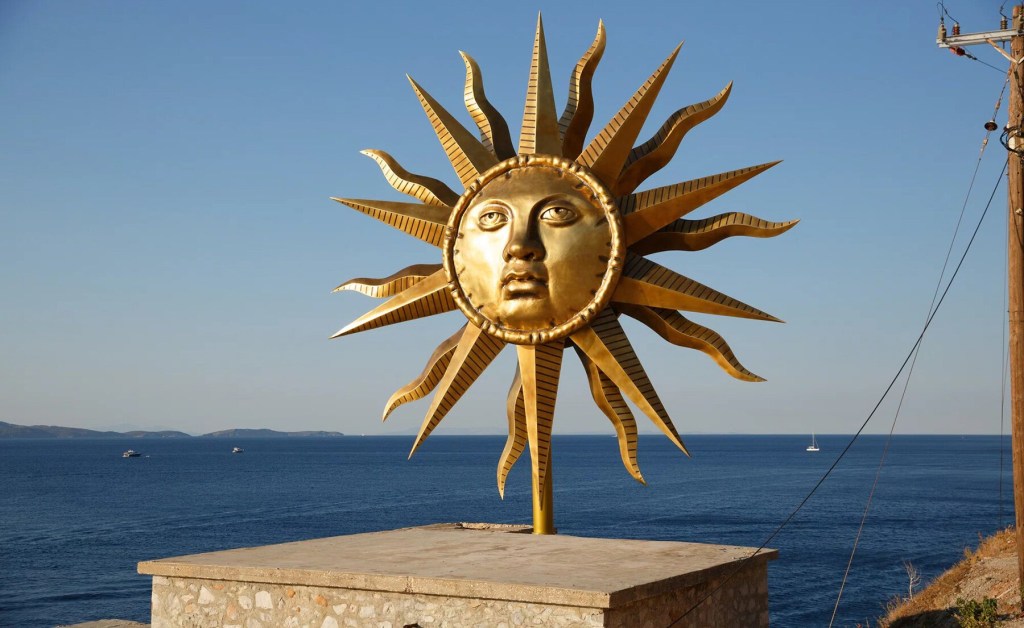 Escultura de bronze de sou com rosto de Apollo. Obra está ao ar livre, diante do mar.