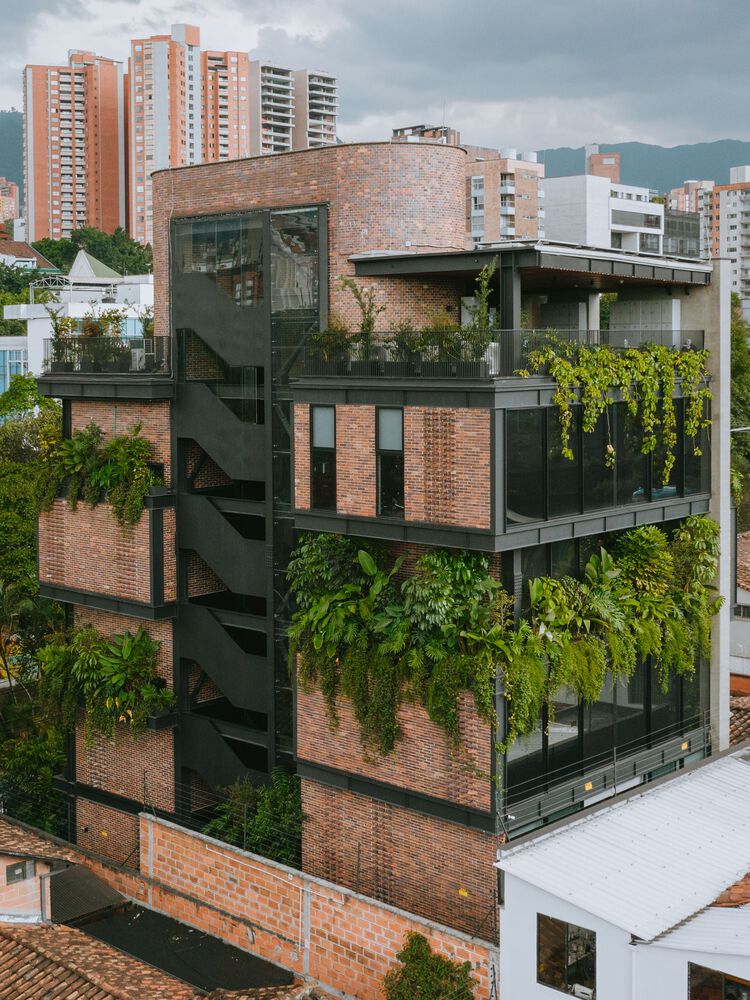 Fachada de prédio com estrutura metálica, tijolinhos aparentes e floreiras com vegetação densa.