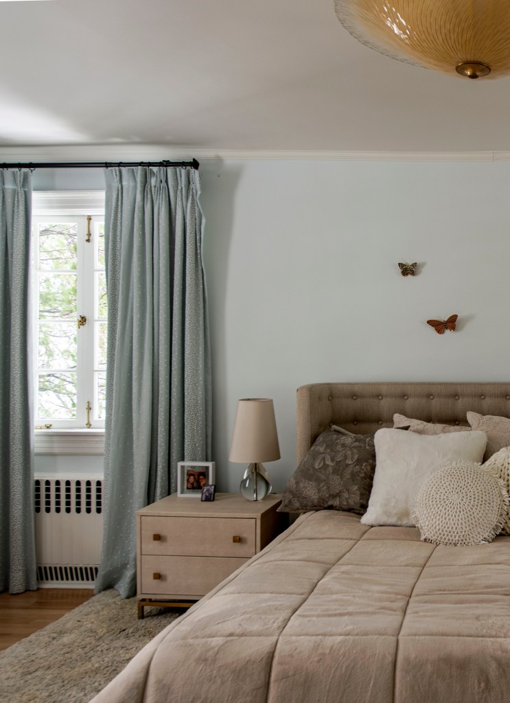 Quarto de casal com cortinas azuis, mesa de cabeceira de madeira e cama com cabeceira. Borboletas na pared