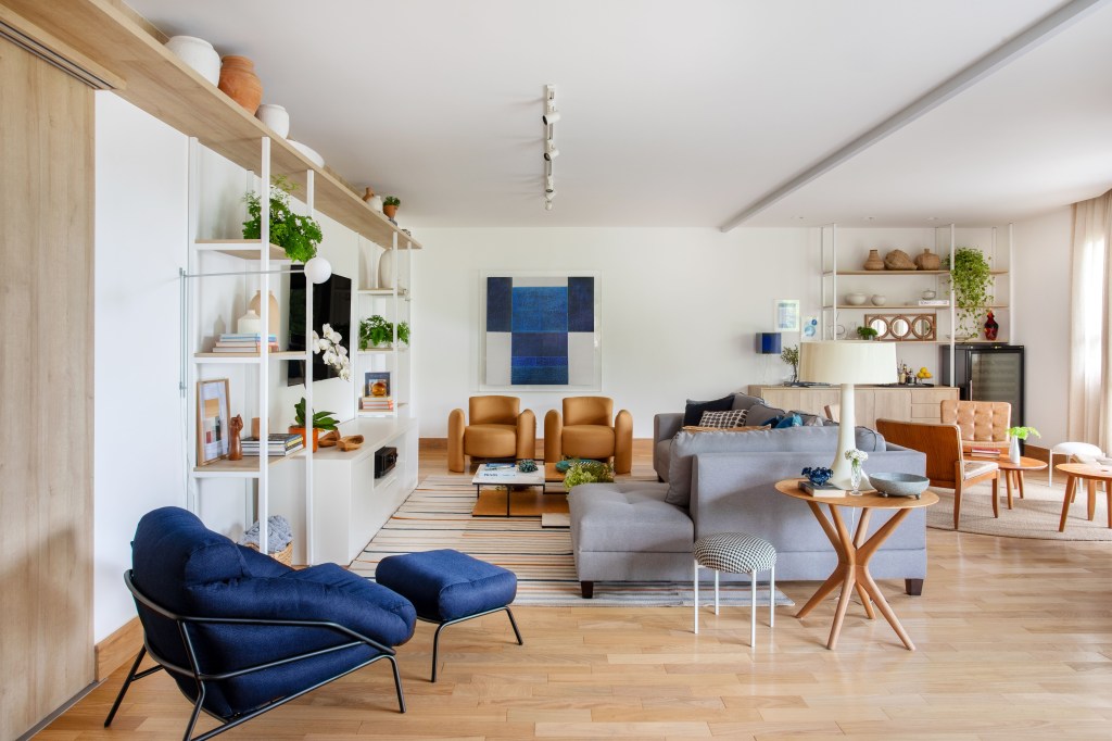 Sala de estar clara com piso em madeira, estante branca, poltrona azul, sofá cinza.