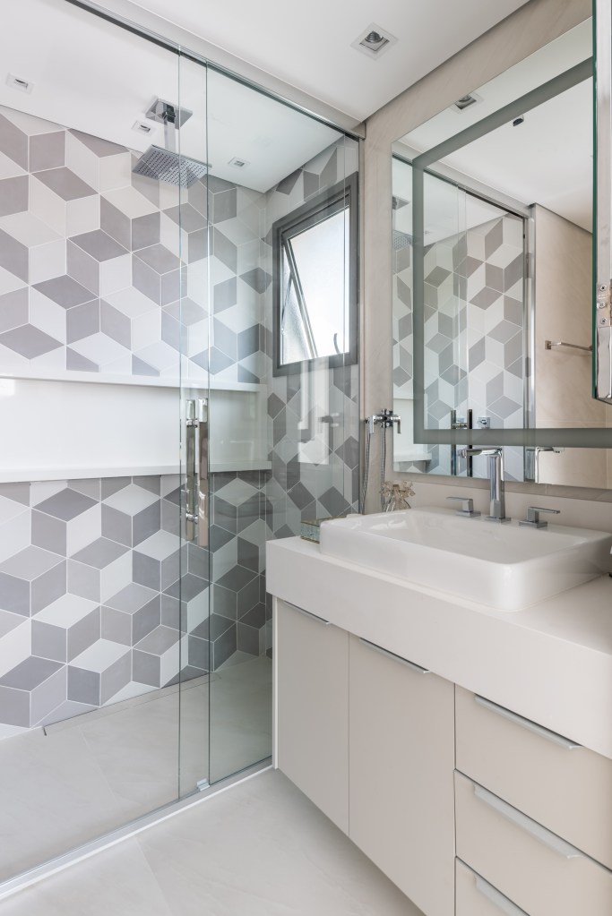 Banheiro com box de vidro e parede de azulejos geométricos brancos e cinzas.
