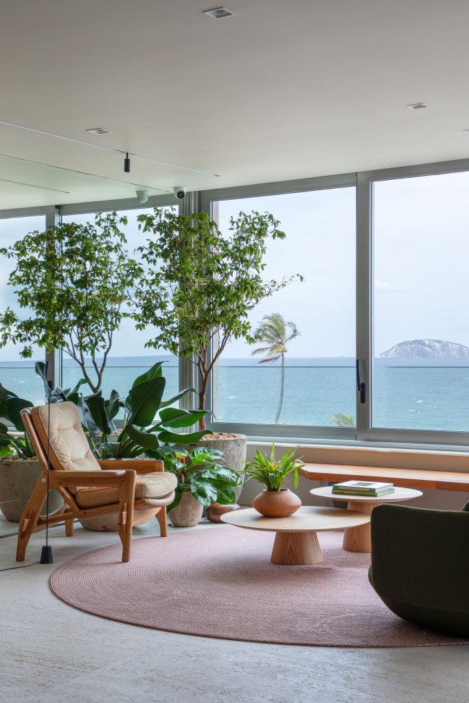 Sala de estar com grandes janelas, tapete redondo rosado, poltrona e plantas.