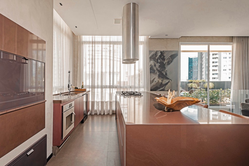 Grandes áreas envidraçadas marcam este dúplex de 220 m² em Curitiba. Projeto do Estudio Elmor. Na foto, cozinha com janelas com cortinas, marcenaria terrosa. Ilha com cooktop e vista do jardim.