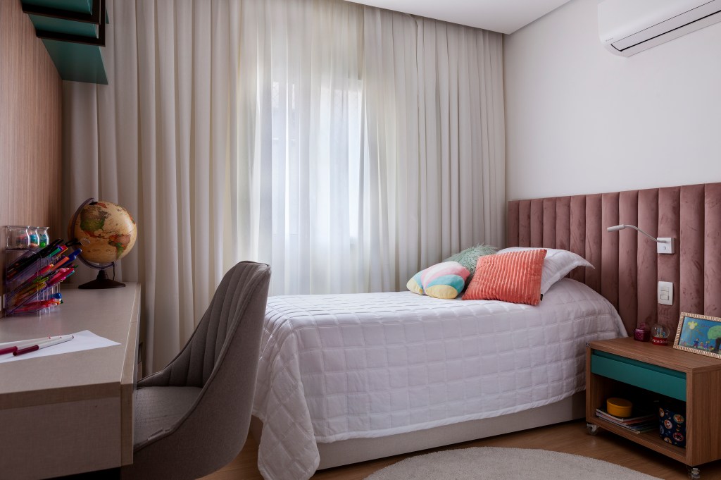 Quarto infantil com cama de solteiro, cabeceira acolchoada, bancada de estudos e cortina.