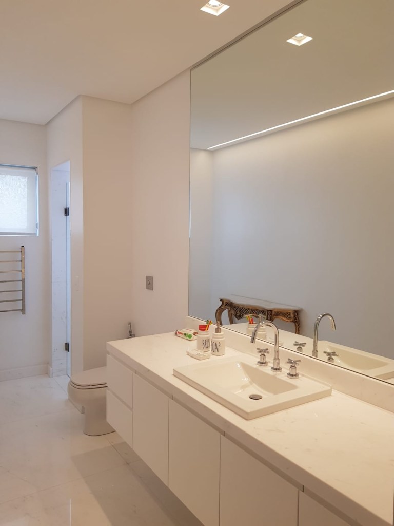 Banheiro branco amplo com bancada branca e espelho grande.