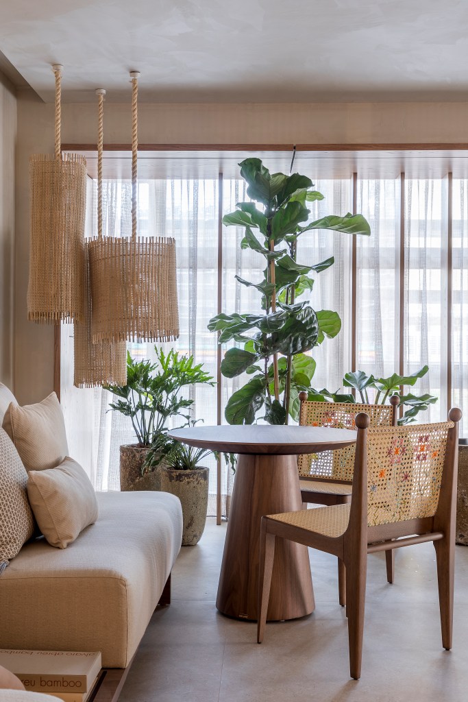 Sala de estar em tons beges, mesa redonda pequena de madeira, cadeiras de palhinha, luminária de palhinha e plantas.