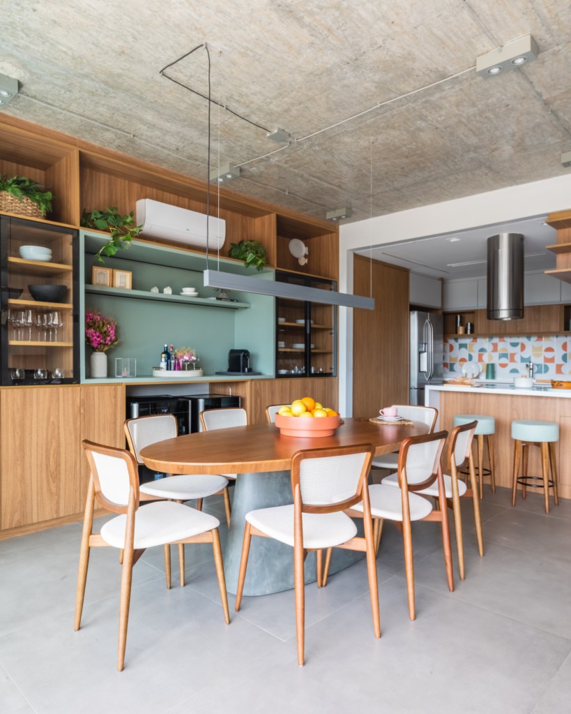 Projeto de BMA Studio. Na foto, sala de jantar integrada com cozinha com mesa oval de madeira, cadeiras brancas e estante de marcenaria com nicho central verde.