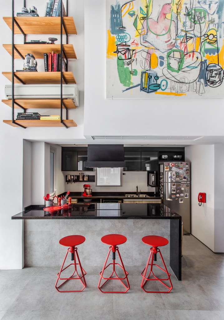 Projeto de Korman Arquitetos. Na foto, cozinha integrada em loft com pé-direito duplo. Bancada preta e banquetas vermelhas.