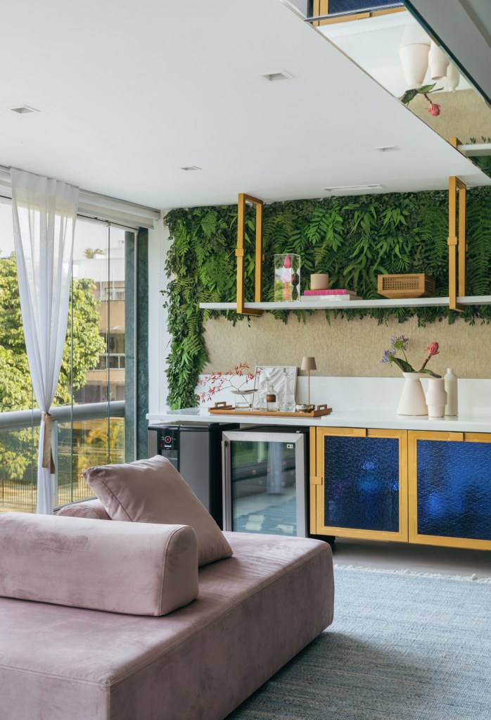 Apê de Ciro Bottini tem paleta Art Deco Navy com tons azuis e dourados. Projeto de Casa Cururu. Na foto, varanda integrada com bar, sofá ilha rosa e jardim vertical.